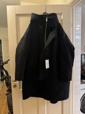 Svart Liam Melton Wool Parka Whyred - Snygg svart parkas från Whyred i modellen Liam Melton Wool Parka. Jackan är tillverkad i ullblandning och har hög krage, dragkedja och knappar framtill. Perfekt för kalla dagar när du vill ha både stil och värme.