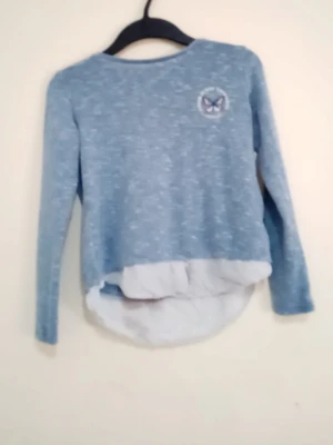 Blå tröja med fjärilstryck - Blåmelerad långärmad tröja med rund hals och dekorativt fjärilstryck på bröstet. Texten runt fjärilen lyder 'Think Sunny Thoughts'. Tröjan har en vit, skjortliknande nederdel som ger en lager-på-lager-look. Perfekt för dig som gillar unika detaljer.