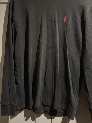 Svart långärmad tröja från Polo Ralph Lauren - Klassisk svart långärmad tröja från Polo Ralph Lauren med den ikoniska röda loggan broderad på bröstet. Tröjan har rund halsringning och är tillverkad i mjukt bomullsmaterial. Perfekt för en stilren och enkel look.   Passar M