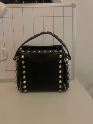 Svart handväska med nitar - Handväska, köpt secondhand (gissar på att den kommer från Zara) i bra skick. Säljer pga att jag sällan använder den. Passar till de mesta outfits. Väskan har dragkedjor på sidorna som används för att justera storleken på väskan.