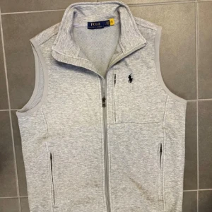 Ljusgrå väst från Polo Ralph Lauren - Sportig ljusgrå väst från Polo Ralph Lauren med dragkedja framtill och tre fickor med dragkedja. Västen har hög krage, meshfoder och klassisk broderad logga på bröstet. Perfekt för lager-på-lager och en clean look.