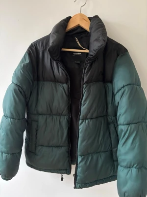 Grön och svart pufferjacka Pull&Bear - Snygg pufferjacka från Pull&Bear i mörkgrönt och svart. Jackan har hög krage, dragkedja framtill och quiltad design. Perfekt för kalla dagar och ger en cool streetwear-look. Tillverkad i syntetmaterial för extra värme.