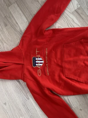Röd hoodie från GANT med tryck - Snygg röd hoodie från GANT med broderad logga och texten 'American Sportswear' på bröstet. Klassisk känguruficka framtill och huva. Tillverkad i mjuk bomull, perfekt för en avslappnad stil.