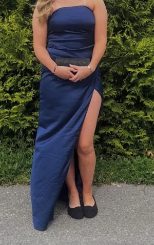 Marinblå långklänning med slits - Köpt för 1000 säljer för 300 Säljer en elegant marinblå långklänning i satinliknande material. Klänningen är axelbandslös och har en hög slits på ena sidan för en snygg och modern look. Perfekt för bal eller festliga tillfällen.