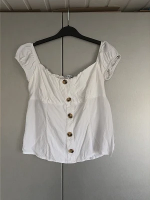 Vit blus med knappar från NA-KD - Supersöt vit blus från NA-KD med offshoulder-modell och korta puffärmar. Blusen har snygga träfärgade knappar framtill och en lätt croppad passform. Perfekt att styla med jeans eller kjol för en fräsch look.