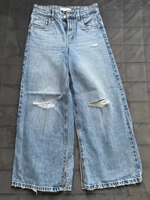 Baggy slitna jeans  - Säljer ett par ljusblå jeans med breda ben och hög midja. Jeansen har slitningar på båda benen och råa kanter vid bensluten för en trendig look. Klassisk femficksmodell i denim. Perfekta för dig som gillar avslappnad stil.