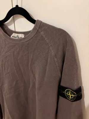 Grå/brun crewneck tröja från Stone Island, size S - Säljer en grå långärmad sweatshirt från Stone Island med klassisk rund hals och den ikoniska logotypen på vänster ärm. Tröjan har ribbade muddar vid ärmslut och nederkant. Perfekt för en avslappnad och stilren look.