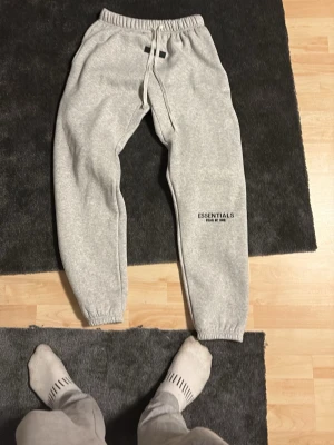 Grå mjukisbyxor från Essentials - Snygga grå mjukisbyxor från Essentials med svart logga på vänster ben. Byxorna har resår i midjan med dragsko och muddar vid bensluten. Perfekta för en chill och avslappnad stil.