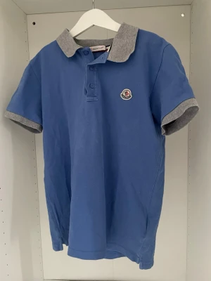 Moncler pike  - Snygg blå pikétröja från Moncler med grå krage och gråa ärmslut. Klassisk polokrage med knappar och Moncler-logga på bröstet. Perfekt för en stilren och avslappnad look.