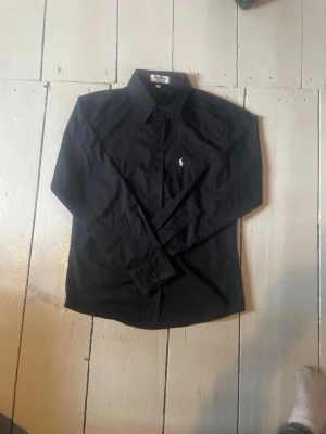 Svart skjorta från Ralph Lauren - Klassisk svart skjorta från Ralph Lauren med vit broderad logga på bröstet. Skjortan har lång ärm, knappar framtill och en stilren krage. Perfekt för dig som gillar en clean och tidlös look.