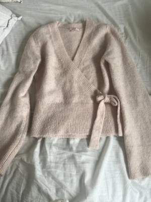 Ljusrosa omlottkofta från Gina Tricot - Mysig ljusrosa stickad omlottkofta från Gina Tricot med knytband i sidan och ribbstickade detaljer vid nederkant och ärmslut. Perfekt för lager-på-lager och har en mjuk, fluffig känsla. Långärmad och med djup v-ringning.