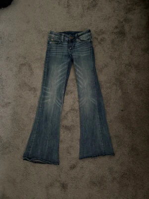 Blå bootcut jeans med slitningar - Säljer ett par blå bootcut jeans med snygga slitningar framtill och klassisk femficksmodell. Jeansen har låg midja och är tillverkade i ett mjukt denimtyg med lätt stretch. Perfekta för en avslappnad och trendig look.