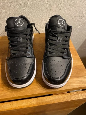 Svarta Air Jordan 1 Low sneakers - Snygga svarta Air Jordan 1 Low sneakers med vit sula och klassisk Jumpman-logga på plösen. Skorna har svart läder och textil, svarta skosnören och en vit kontrastsöm på Swoosh-loggan. Perfekta för dig som gillar streetwear och vill ha en ikonisk look.