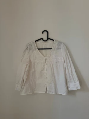 Vit blus med spetsdetaljer - Söt vit blus med v-ringning och knappar framtill. Blusen har fina spetsdetaljer längs bröstet och ärmarna samt lätt puffade trekvartsärmar. 