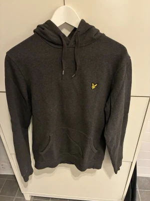Mörkgrå hoodie från Lyle & Scott - Mörkgrå hoodie från Lyle & Scott med klassisk gul logga på bröstet. Hoodien har dragsko i huvan och en stor känguruficka framtill. Tillverkad i mjukt bomullsmaterial, perfekt för en avslappnad stil.