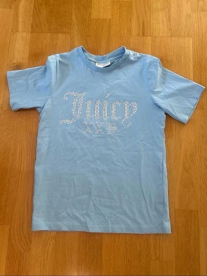 Blå Juicy Couture t-shirt med strass - Ljusblå t-shirt från Juicy Couture med korta ärmar och rund hals. Framsidan pryds av texten 'Juicy XXXY' i glittrande strass. Perfekt för dig som gillar en trendig och glamorös look. Tillverkad i mjuk bomull för skön känsla.