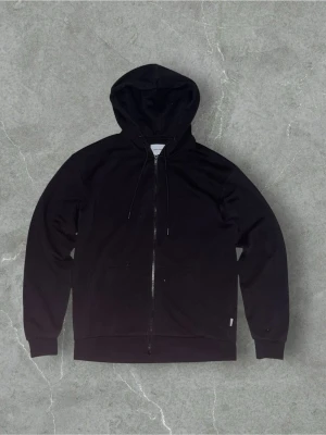 Jack & jones zip hoodie - Klassisk svart hoodie med dragkedja framtill och justerbar huva. Tillverkad i mjukt bomullsmaterial och har två fickor på magen. Perfekt för en avslappnad och stilren look. Storlek S.