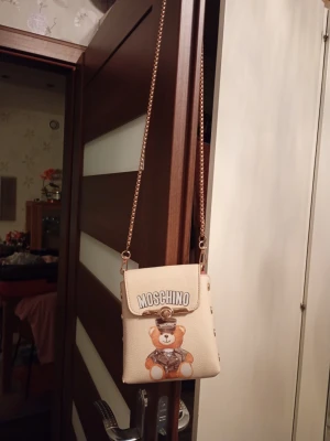 Beige Moschino handväska med kedja - Söt beige handväska från Moschino med guldfärgad kedjerem och ett tryck av en nallebjörn på framsidan. Väskan har ett lock med vridlås och röd insida. Läder.