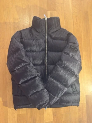 Svart pufferjacka från Dior - Snygg svart pufferjacka från Dior med diskret monogrammönster över hela jackan och hög krage. Jackan har en silverfärgad dragkedja med Dior-logga och patch med Dior på ärmen. Perfekt för kalla dagar och ger en exklusiv look.