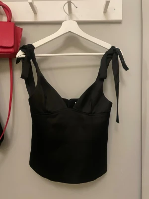 Svart korsettopp med knytband - Stilren svart korsettopp med breda axelband som knyts upptill. Toppen har en figurnära passform och är tillverkad i ett glansigt material som ger en lyxig känsla. Perfekt att styla med jeans eller kjol för en trendig look.