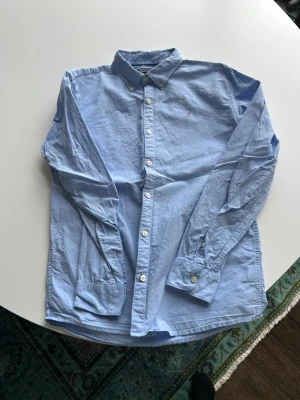 Ljusblå skjorta från Tommy Hilfiger stl 176  - Klassisk ljusblå skjorta från Tommy Hilfiger med button-down krage och vita knappar. Skjortan har lång ärm och den ikoniska lilla loggan broderad på bröstet. Tillverkad i mjuk bomull för en clean och stilren look. Obs liten i storleken! 