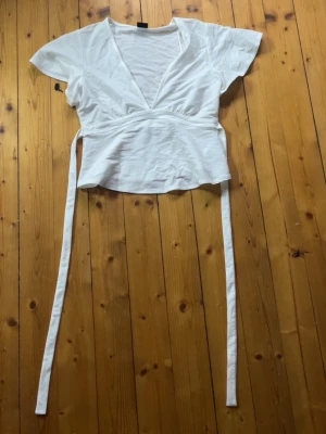 Vit tie topp med knytband - Säljer en vit omlottopp med v-ringning i storlek S och knytband i midjan. Toppen har korta ärmar och ganska tunt material. Superfin verkligen men passar inte så bra på mig. Aldrig använt så helt ny utan prislapp!💕