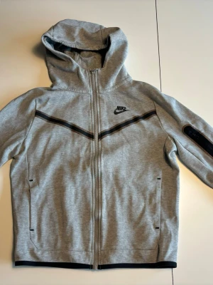Grå Nike hoodie med dragkedja - Snygg grå hoodie från Nike med hel dragkedja och svart Nike-logga på bröstet. Hoodien har svarta detaljer i form av ränder över bröstet och på ena ärmen. Utrustad med huva och praktiska fickor framtill. Perfekt för en sportig och avslappnad stil.