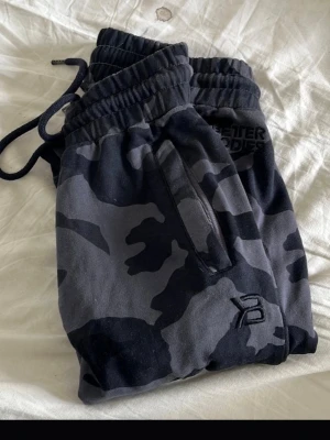 Camo mjukisbyxor från Better Bodies - Säljer ett par snygga camofärgade mjukisbyxor från Better Bodies. Byxorna har svart och grått camomönster, dragsko i midjan och dragkedjeförsedda fickor. Perfekta för chill eller gymmet, med broderad logga på benet.