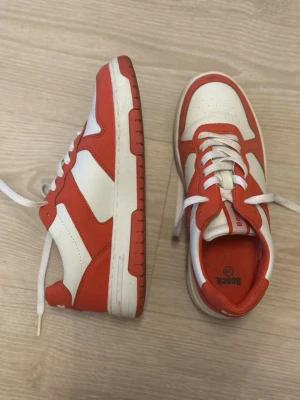 Rödvita sneakers från Bench - Snygga sneakers från Bench i rött och vitt med klassisk retrostil. Skorna har vita snören, perforerad tåbox och kontrasterande röda detaljer. Ovandelen är i syntetmaterial och sulan är vit med röda inslag. Perfekta för dig som gillar streetwear och sportig look. Använt max 5 ggr
