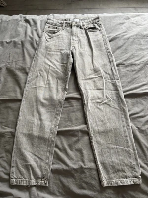 Ljusgrå raka jeans - Säljer ett par ljusgrå jeans med rak passform och klassisk femficksdesign. Jeansen har normal midja och är tillverkade i denim. Perfekta för en avslappnad och trendig stil. Något slitet längs ner men själv tycker det ser coolare ut.