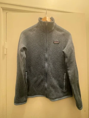 fleecejacka från Patagonia - Mysig grå fleecejacka från Patagonia med hel dragkedja och hög krage. Två fickor framtill med dragkedja och diskret logga på bröstet. Perfekt för lager-på-lager och outdoor-vibe. Materialet är mjukt och värmande.