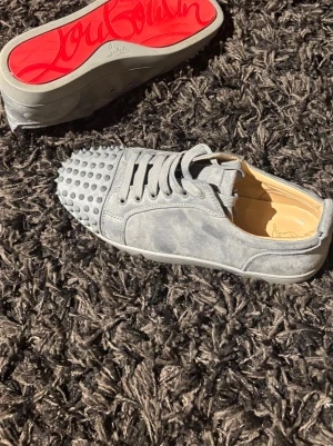 Gråa Louboutin sneakers med nitar - Unika grå sneakers från Christian Louboutin i mocka med ikoniska röda sulor och coola nitar på tån. Skorna har lågt skaft, matchande grå skosnören och en lyxig känsla som sticker ut. Perfekta för dig som vill ha något extra till din outfit.