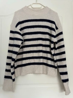 Randig stickad tröja i beige och marinblå - Säljer en ribbstickad tröja med breda marinblå ränder på beige botten. Tröjan har rund halsringning, långa ärmar och avslappnad passform. Perfekt för dig som gillar klassisk och enkel stil. Mjuk i materialet, använd några gånger men fortfarande fin! 