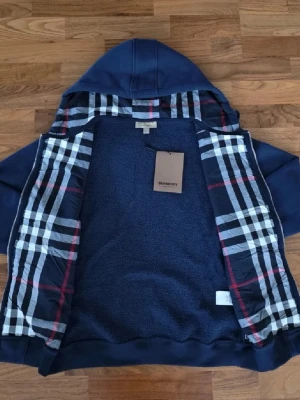 Marinblå hoodie från Burberry - Snygg marinblå hoodie från Burberry med dragkedja och klassiskt rutigt foder i huvan och insidan. Hoodien har fickor framtill, ribbade muddar och justerbar huva med snören. Perfekt för dig som vill ha en stilren och exklusiv look.