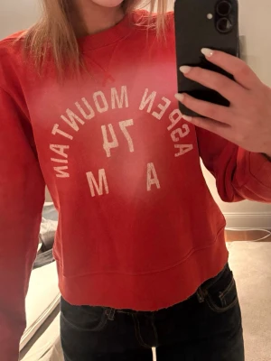 Röd sweatshirt Aspen Mountain - Cool röd sweatshirt med vit text 'Aspen Mountain 1974 Rugby U.S.A Western Colorado' både fram och bak. Tröjan har rund hals, långa ärmar och ribbade muddar. Perfekt för en avslappnad och sportig look.