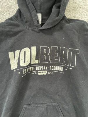 Svart Volbeat hoodie med tryck - Säljer en svart hoodie från Volbeat med stort tryck på bröstet och en stor bild på ryggen. Texten 'Rewind • Replay • Rebound' syns tydligt. Hoodien har huva och magficka framtill. Perfekt för dig som gillar bandmerch och vill ha en cool streetstil. Den har en fläck på ryggen och ena ärmen men är lätt att ta bort med rätt medel. Storleken står inte men den är i storlek S men skulle säga att den är mer i XS.