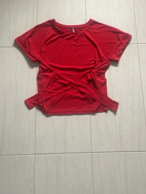 Knyt top från jyothi  - Cool röd topp med omlott-design och knytband framtill. Kortärmad och croppad modell som ger en snygg och trendig look. Perfekt för dig som vill sticka ut med färg och stil. Materialet känns mjukt och lätt, troligen polyester.