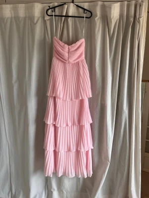 Rosa volangklänning med plissering - Söt rosa klänning med flera lager volanger och plisserat tyg. Axelbandslös modell med dragkedja bak. Den är perfekt till bal eller annan finare tillställning då det behövs en vacker långklänning.