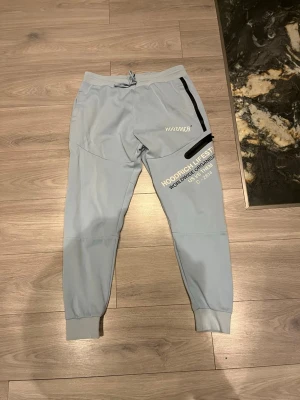 Ljusblå mjukisbyxor från Hoodrich - Snygga ljusblå mjukisbyxor från Hoodrich med tryck på benet och svarta detaljer vid fickorna. Byxorna har resår i midjan med dragsko och muddar vid bensluten. Perfekta för en avslappnad streetwear-look.