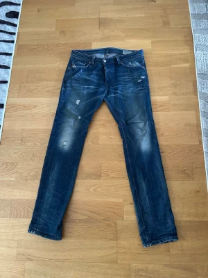 Blå slitna jeans från Diesel - Säljer ett par blå jeans från Diesel med snygga slitningar och distressed detaljer. Jeansen har klassisk femficksmodell, raka ben och tydliga tvättade partier för en cool, avslappnad look. Perfekta för dig som gillar streetwear och en trendig stil.