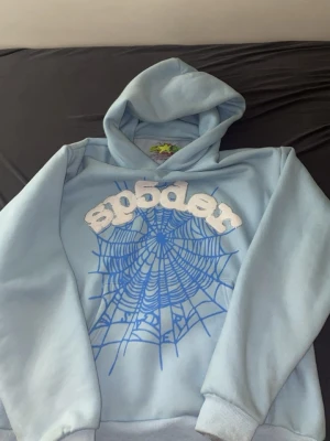 Ljusblå hoodie med sp5der-tryck - Ljusblå hoodie från Sp5der med stor huva och ribbade muddar. Framsidan har ett stort blått spindelnätsmönster och texten 'sp5der' i vitt med glittriga detaljer. Tillverkad i mjukt material, perfekt för en avslappnad streetwear-look.