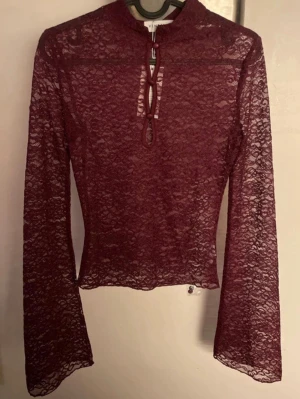 Vinröd spetsblus med knyt - Vinröd långärmad topp i genombruten spets med knappar vid halsen. Figurnära modell med lätt utsvängda ärmar och hög hals. Perfekt för dig som gillar trendiga och feminina detaljer.