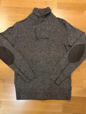 Grå stickad tröja med dragkedja - Mörkgrå stickad tröja med hög krage och kort dragkedja framtill. Tröjan har förstärkta armbågslappar i mörkare tyg och ribbade muddar vid ärmslut och nederkant. Perfekt för kyliga dagar och en avslappnad stil.