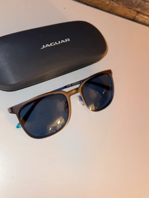 solglasögon från Jaguar - Snygga solglasögon från Jaguar med gråa bågar och blått glas. Klassisk rektangulär form och diskret logga på skalmen. Kommer med originalfodral och putsduk. Perfekta för soliga dagar och ger en stilren look. Aldrig använda eftersom det kom dubbelt av dom.