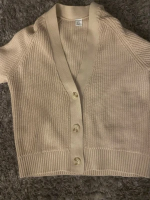 Beige ribbad kofta med knappar - Kort beige kofta med v-ringning och tre stora knappar framtill. Koftan har ribbade muddar och är tillverkad i ett mjukt, stickat material. Perfekt för lager på lager och enkel att matcha med olika outfits.