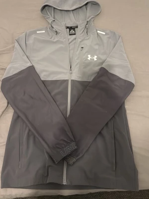 Under Armour windbreaker - Snygg tvåtonad grå vindjacka från Under Armour med huva och hel dragkedja framtill. Jackan har långa ärmar, två sidofickor och logga på bröstet. Perfekt för dig som vill ha en sportig och stilren look. Jackan är unik, Säljs inte längre på nättet, den är i mycket bra skick och är använd fåtal gånger, inga konstigheter med den, kom dm för frågor. 