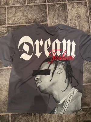 Travis scott merch, t shirt  - Cool grå t-shirt med stort Dream Fashion-tryck på ryggen och mindre Dream-tryck på bröstet. Motiv av en person med kedja på baksidan. T-shirten har rund hals och korta ärmar, perfekt för dig som gillar streetstyle och grafiska prints. Travis t shirt