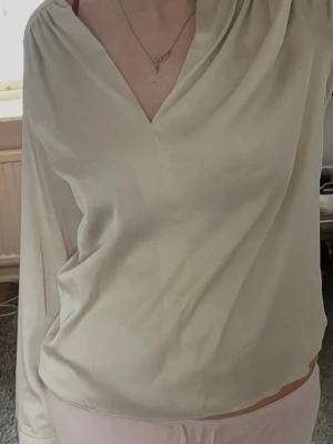 Krämvit blus med v-ringning - Snygg krämvit blus i ett glansigt, mjukt material. Blusen har v-ringning och långa ärmar med lätt drapering vid axlarna. Perfekt för en stilren och enkel look. 