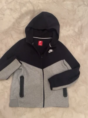 Nike tvåfärgad hoodie med dragkedja - Nike hoodie i svart och grått med dragkedja framtill och två fickor med svarta detaljer. Klassisk huva och Nike-logga broderad på bröstet. Tillverkad i mjukt material som känns skönt mot huden. 