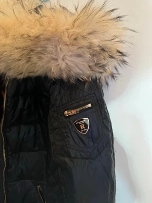 Svart parkas med päls från Rockandblue - Snygg svart parkas från Rockandblue med glansigt material, guldiga dragkedjor och stor huva med fluffig äkta päls. Jackan har quiltad design och en cool logga på ärmen. Perfekt för kalla dagar när du vill vara varm.